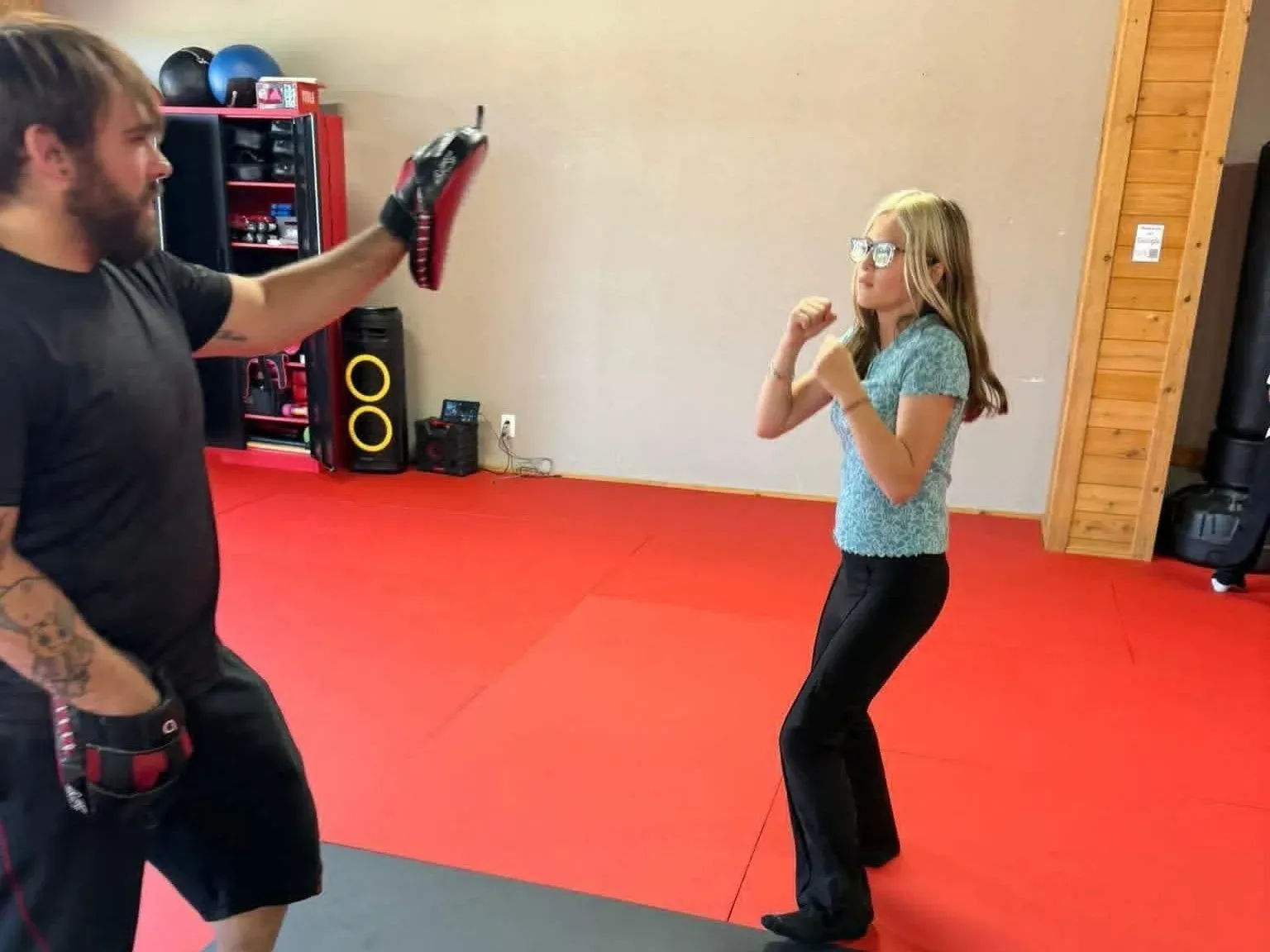A Davis Martial Arts coach holding a focus mitt while a camp kid in street clothes sets her stance on the red mats in Pilot Point, TX