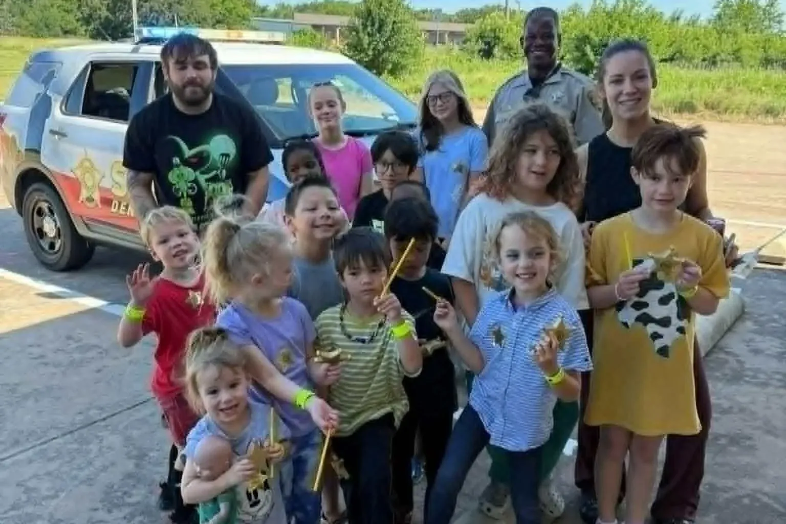 Summer camp kids holding junior deputy badges beside a Denton County Sheriff patrol car during a public-safety visit at Davis Martial Arts & Fitness in Pilot Point, TX