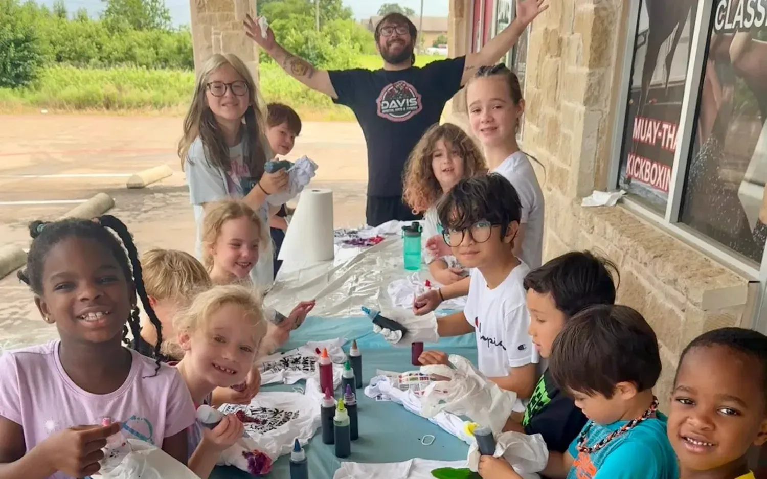 A Davis Martial Arts coach with his arms up behind a long tie-dye table surrounded by summer camp kids decorating their camp shirts in Pilot Point, TX