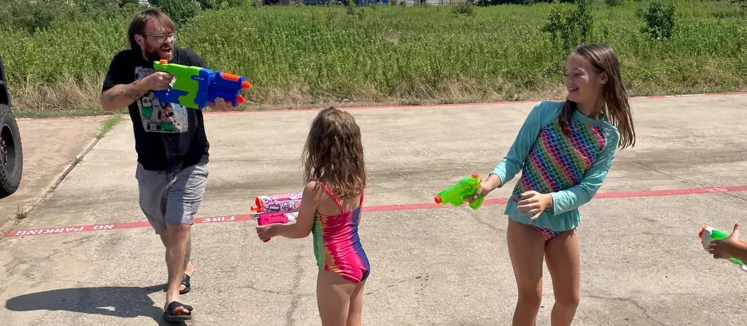 A coach and two summer camp kids in swimsuits trading water-gun fire in the lot behind Davis Martial Arts & Fitness in Pilot Point, TX