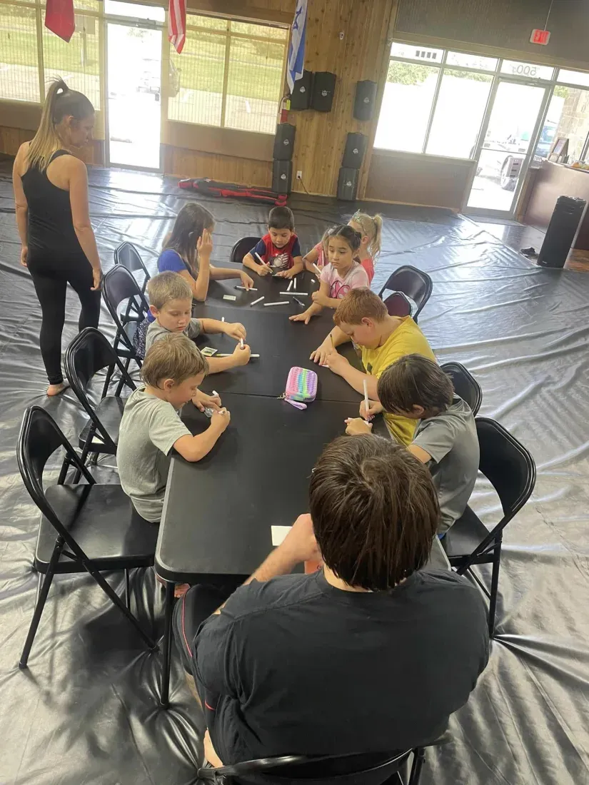 Summer camp kids seated around folding tables doing crafts with a coach standing over them inside Davis Martial Arts & Fitness