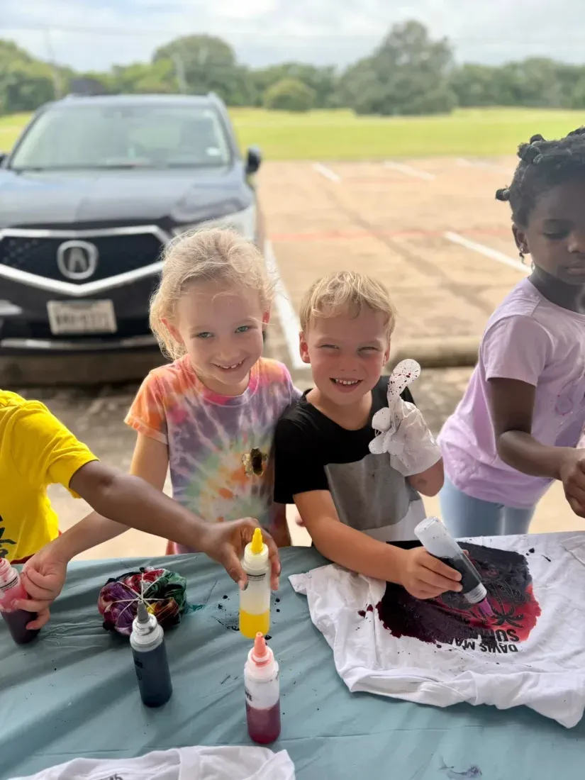 Three summer camp kids squeezing dye bottles onto white shirts spread across a covered table at Davis Martial Arts & Fitness