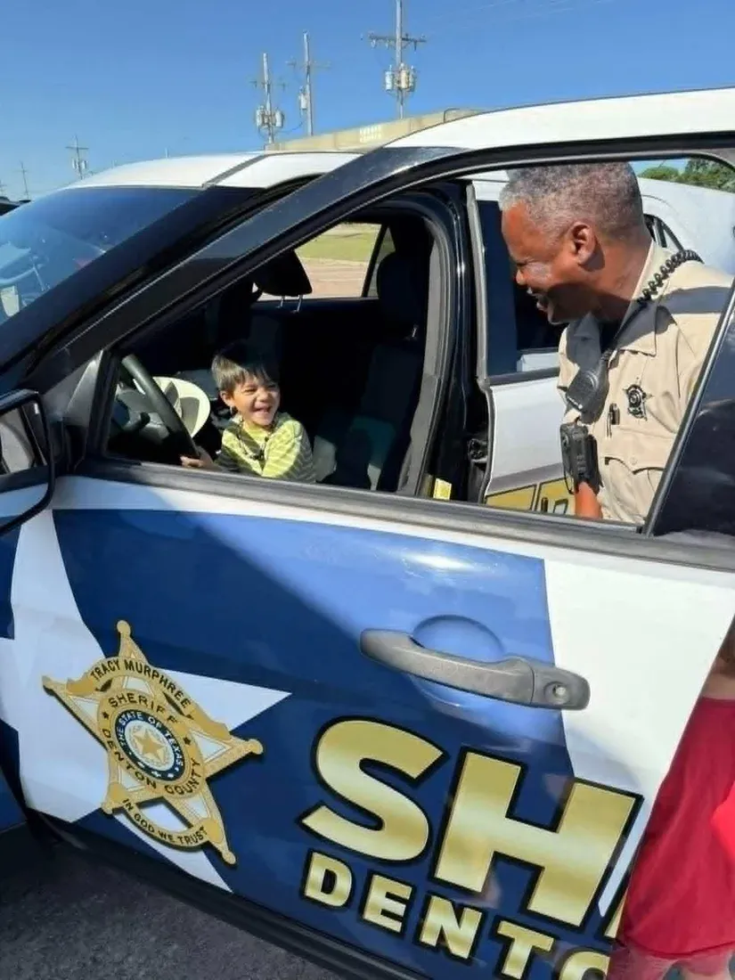 A summer camper sitting behind the wheel of a Denton County Sheriff patrol car while a deputy leans in and laughs, outside Davis Martial Arts & Fitness