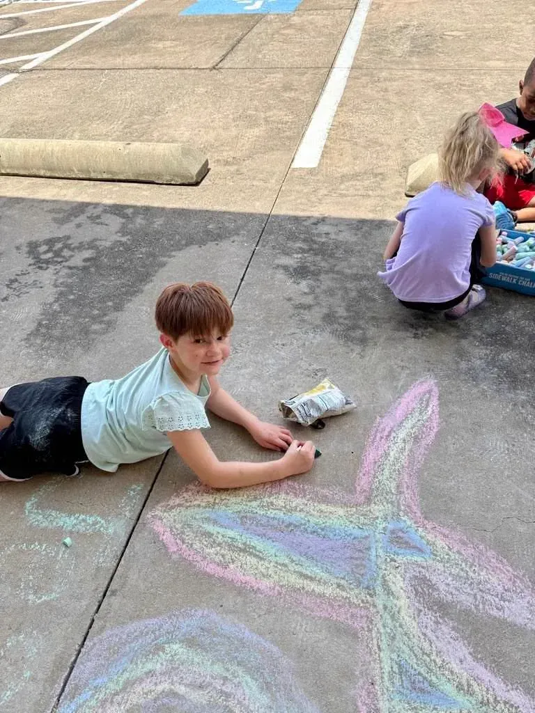 A summer camper sitting on the pavement drawing a large chalk flower outside Davis Martial Arts & Fitness in Pilot Point, TX