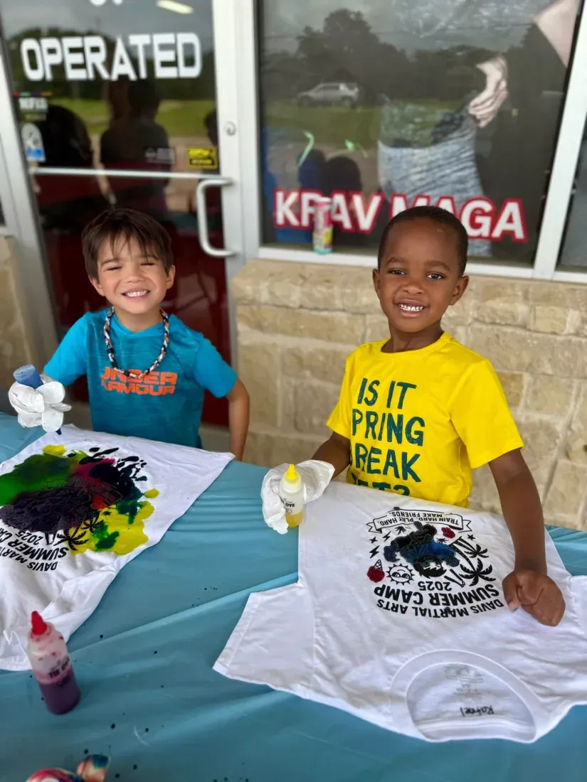Two summer camp boys holding up the Davis Martial Arts Summer Camp shirts they just tie-dyed, outside the academy in Pilot Point, TX