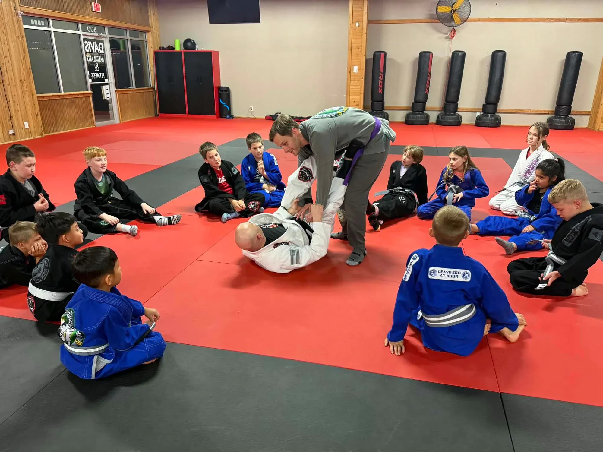 Kids in gis seated around the mat watching a coach demonstrate a technique on a partner during Brazilian Jiu-Jitsu class at Davis Martial Arts & Fitness in Pilot Point