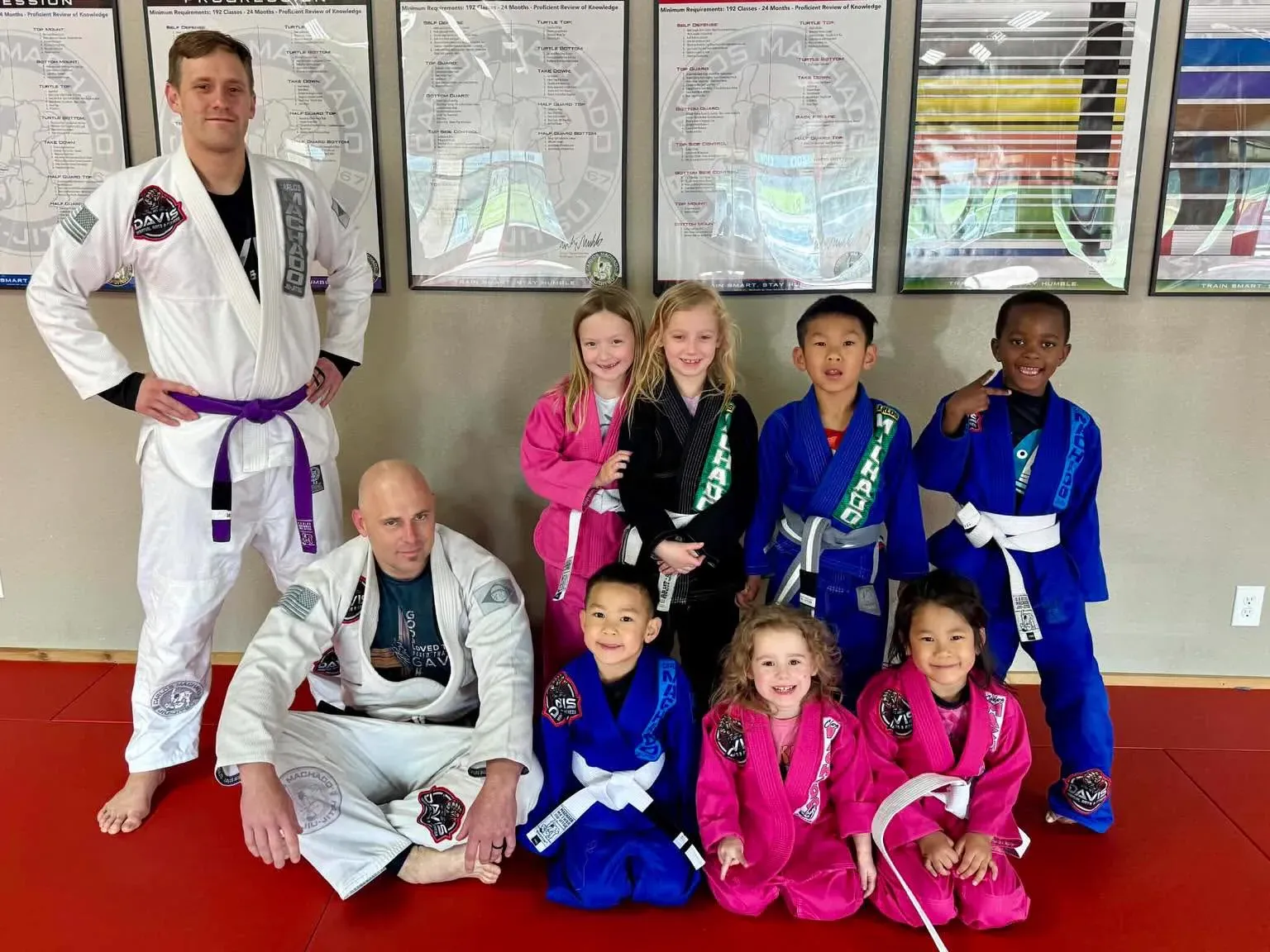 Eight young students in gis smiling with two coaches in front of the Machado curriculum charts at Davis Martial Arts & Fitness in Pilot Point, TX