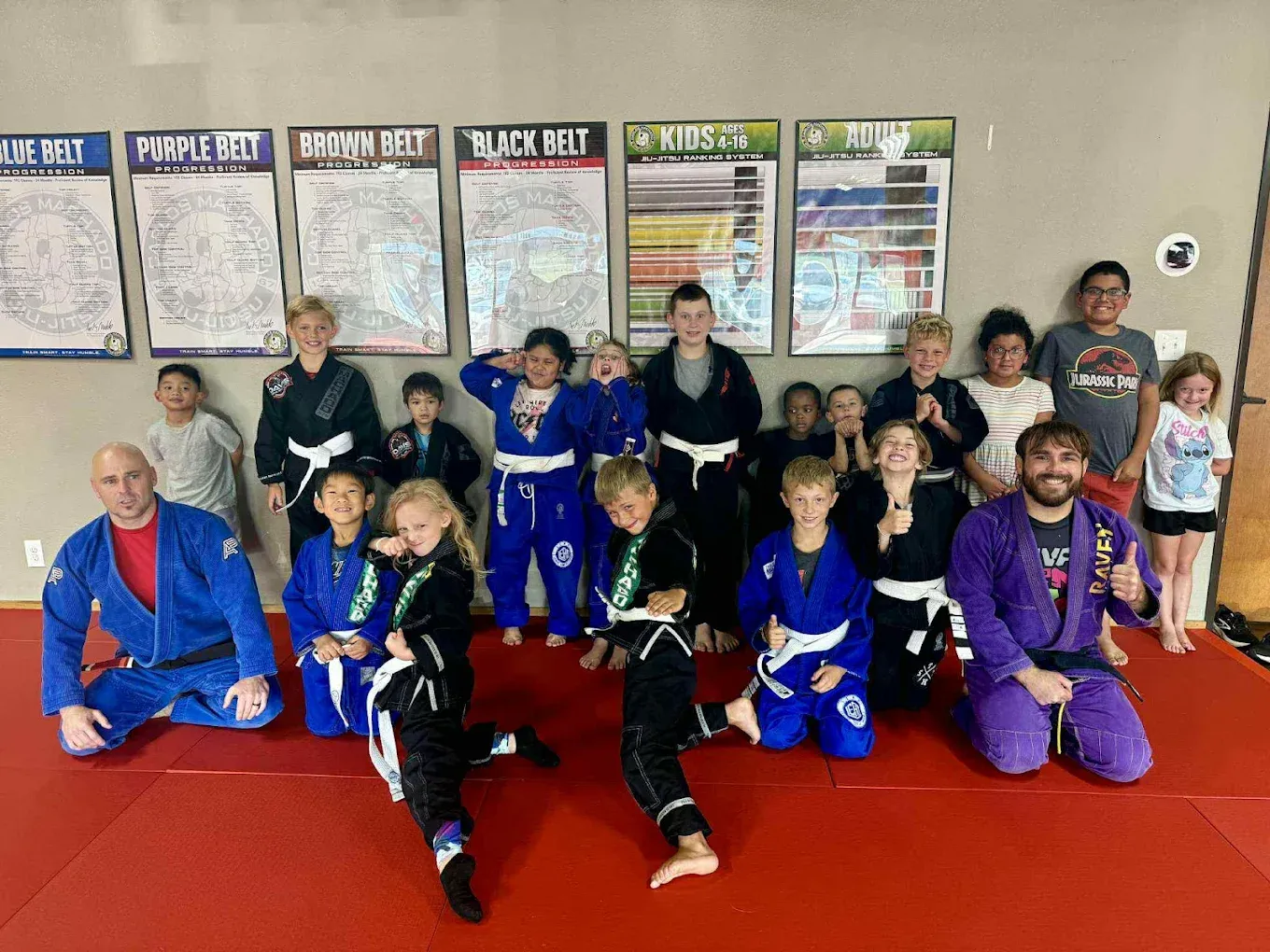 The kids Brazilian Jiu-Jitsu class gathered with two coaches on the mat, belt progression charts on the wall behind them, at Davis Martial Arts & Fitness in Pilot Point, TX