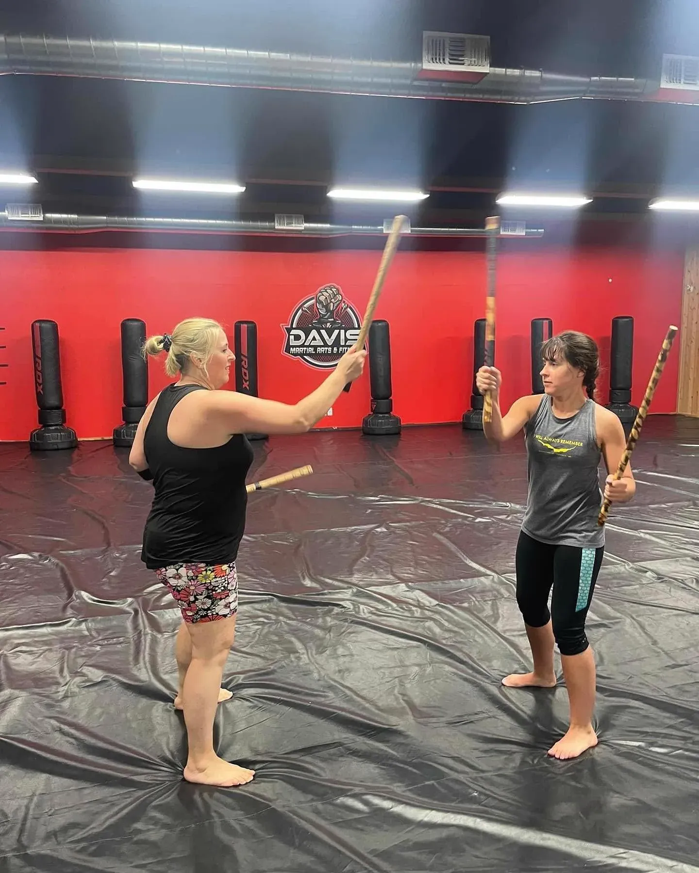 Two women drilling a stick-defense technique together on the mat in front of the Davis logo wall at Davis Martial Arts & Fitness in Pilot Point, TX