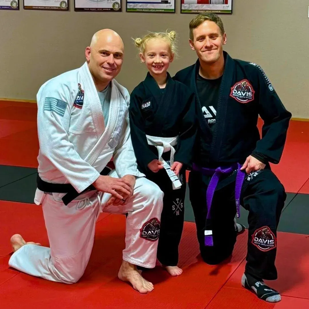 A young student in a black jiu-jitsu gi smiling while holding a new white belt, standing between two Davis Martial Arts coaches on the mat